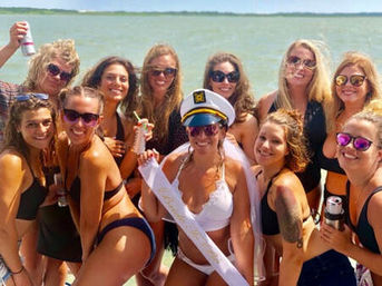 Sunlit group of women in bikinis posing on a boat near the ocean for a bachelorette celebration, bride wearing a captain’s hat, veil and sash, friends smiling and holding drinks.