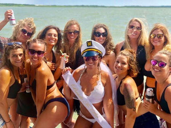 Sunlit group of women in bikinis posing on a boat near the ocean for a bachelorette celebration, bride wearing a captain’s hat, veil and sash, friends smiling and holding drinks.