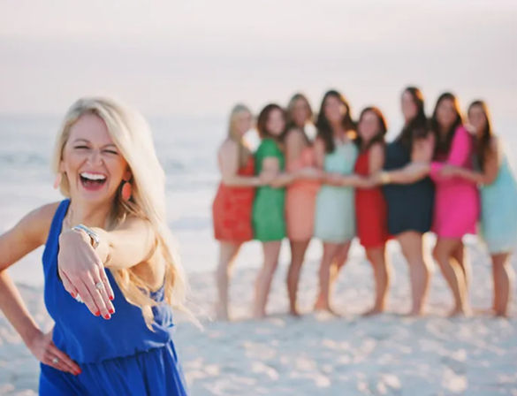 Laughing woman in a blue dress showing her engagement ring on a sunny sandy beach, blurred group of friends in colorful summer dresses linked arm‑in‑arm by the ocean
