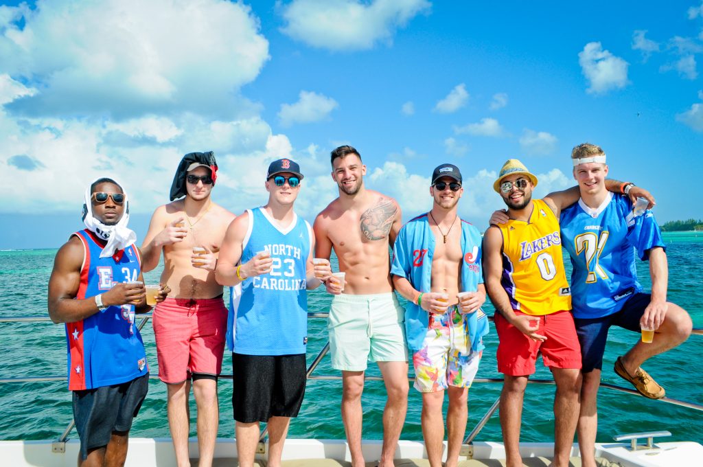 Seven friends in colorful jerseys and swim trunks posing with drinks on a boat over turquoise ocean under a bright blue sky — lively summer yacht party/tropical getaway.