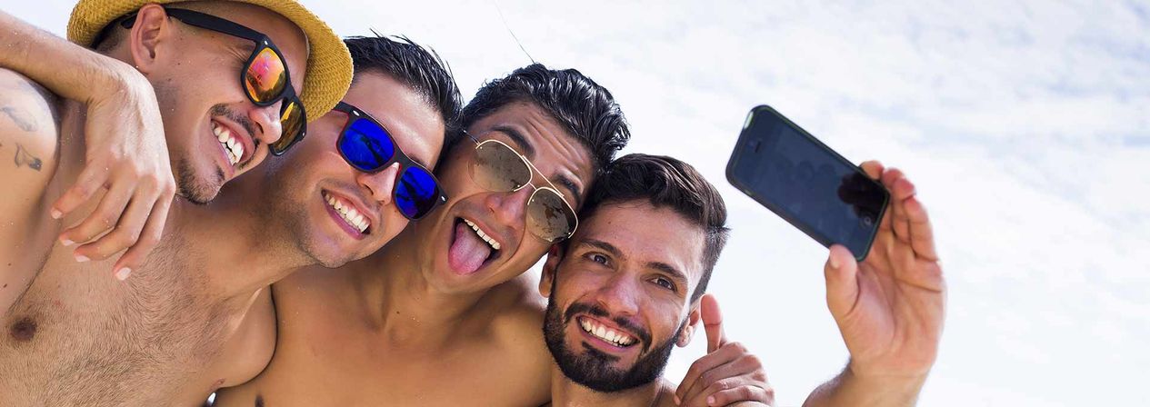 Four friends on a sunny beach taking a joyful selfie, wearing sunglasses and a straw hat against a bright sky