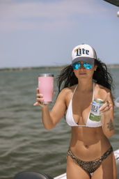 Woman in white bikini and trucker hat with reflective blue sunglasses on a boat over calm bay waters, holding a pink insulated tumbler and a canned drink — sunny summer boating scene.