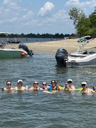 Eight people floating waist-deep in lake water, lined up holding drinks with anchored motorboats and a sandy shoreline under a sunny sky