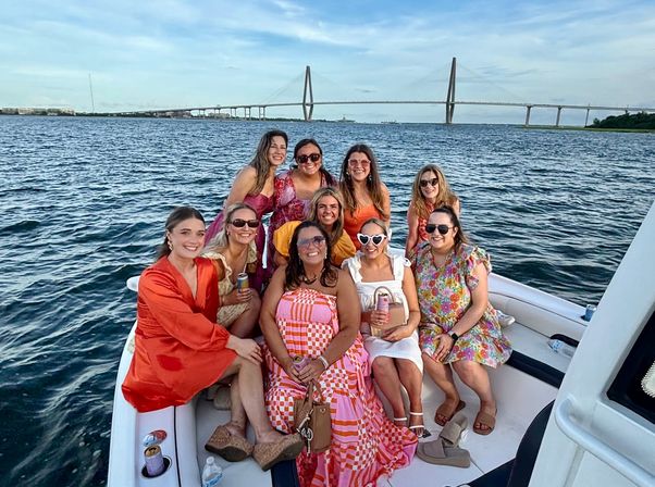 Cheerful group of women in colorful summer dresses on a boat in a coastal harbor, holding drinks with a cable-stayed bridge and blue sky in the background.