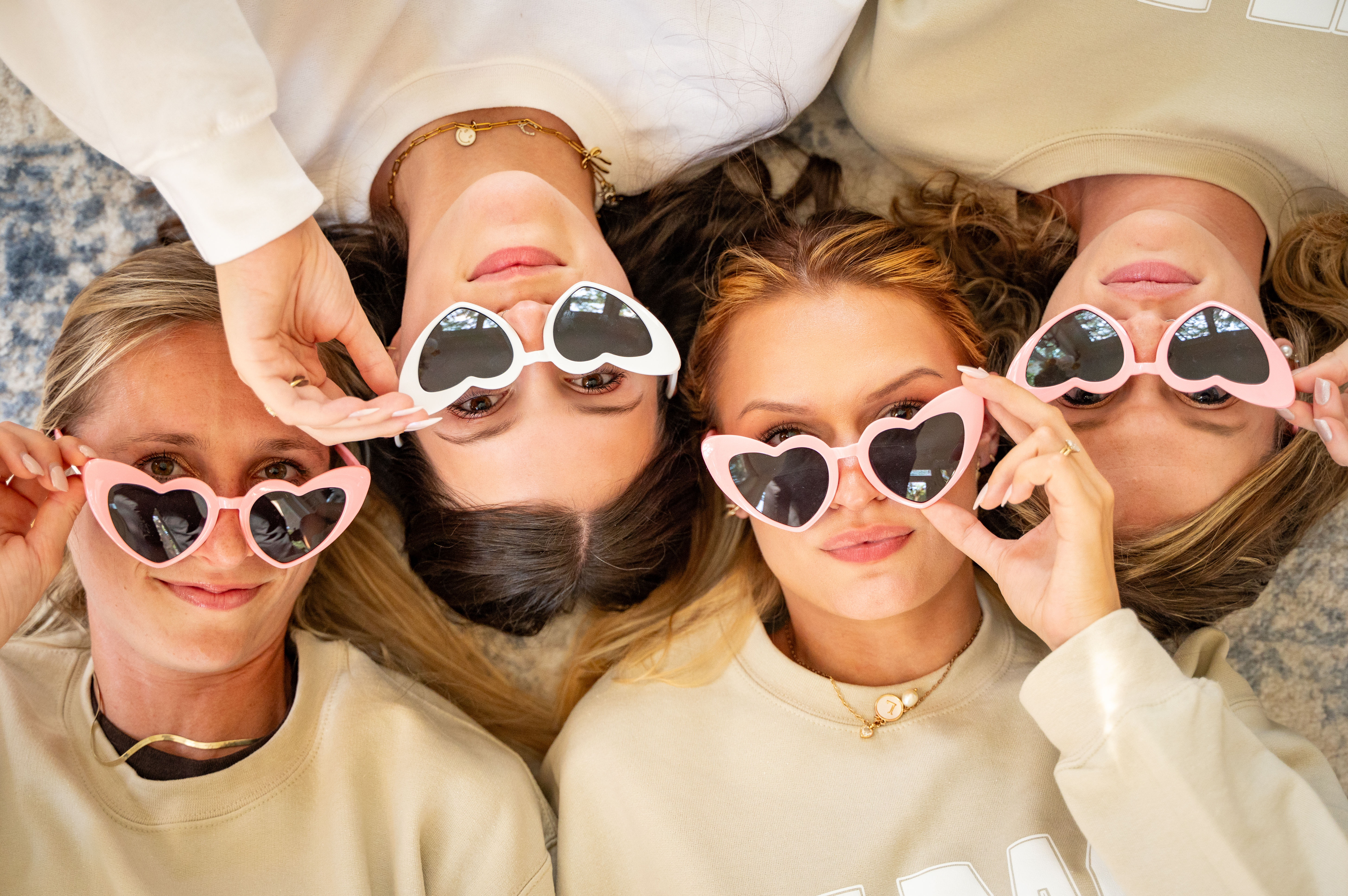 Four women lying head-to-head on a light rug, wearing pink and white heart-shaped sunglasses and cream sweatshirts, playfully posing for an overhead close-up group portrait.