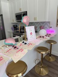 Bright modern white kitchen island styled for a pink seaside bachelorette: gold stools and faucet, shell-shaped balloons, canvas tote favor bags with pink shell prints, heart-shaped sunglasses and a glowing "XOXO" neon sign.