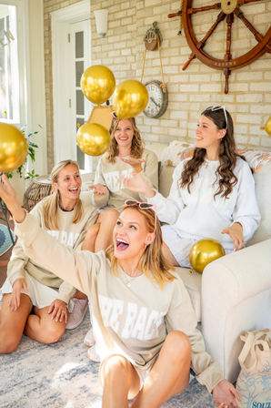Four smiling women in matching beige sweatshirts toss gold balloons in a bright coastal-style sunroom with ship-wheel wall decor — playful indoor celebration.