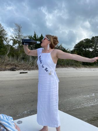 Bride-to-be in a white dress and sash drinking champagne with arms outstretched on a sandy beach shoreline, cloudy coastal sky and treeline in the background