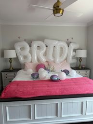 Master bedroom set for a bridal celebration with large white inflatable 'BRIDE' letters over a white bed, pink knit throw, decorative pillows, scattered white and purple balloons, and a gold ceiling fan.
