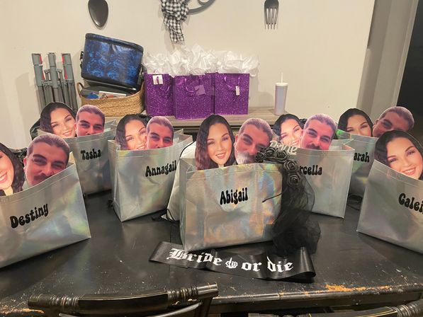 Iridescent personalized gift bags with large cutout face props and visible names on a dining table, purple glitter gift boxes and a “Bride or Die” sash — bachelorette party favors in a home setting.