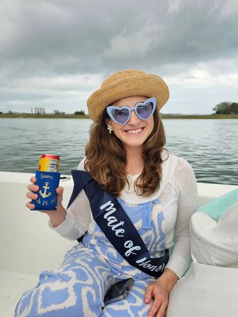 Smiling person on a small boat in a coastal bay, wearing a straw hat, blue heart-shaped sunglasses and a "Mate of Honor" sash, holding a canned drink in a blue anchor koozie under a cloudy sky.