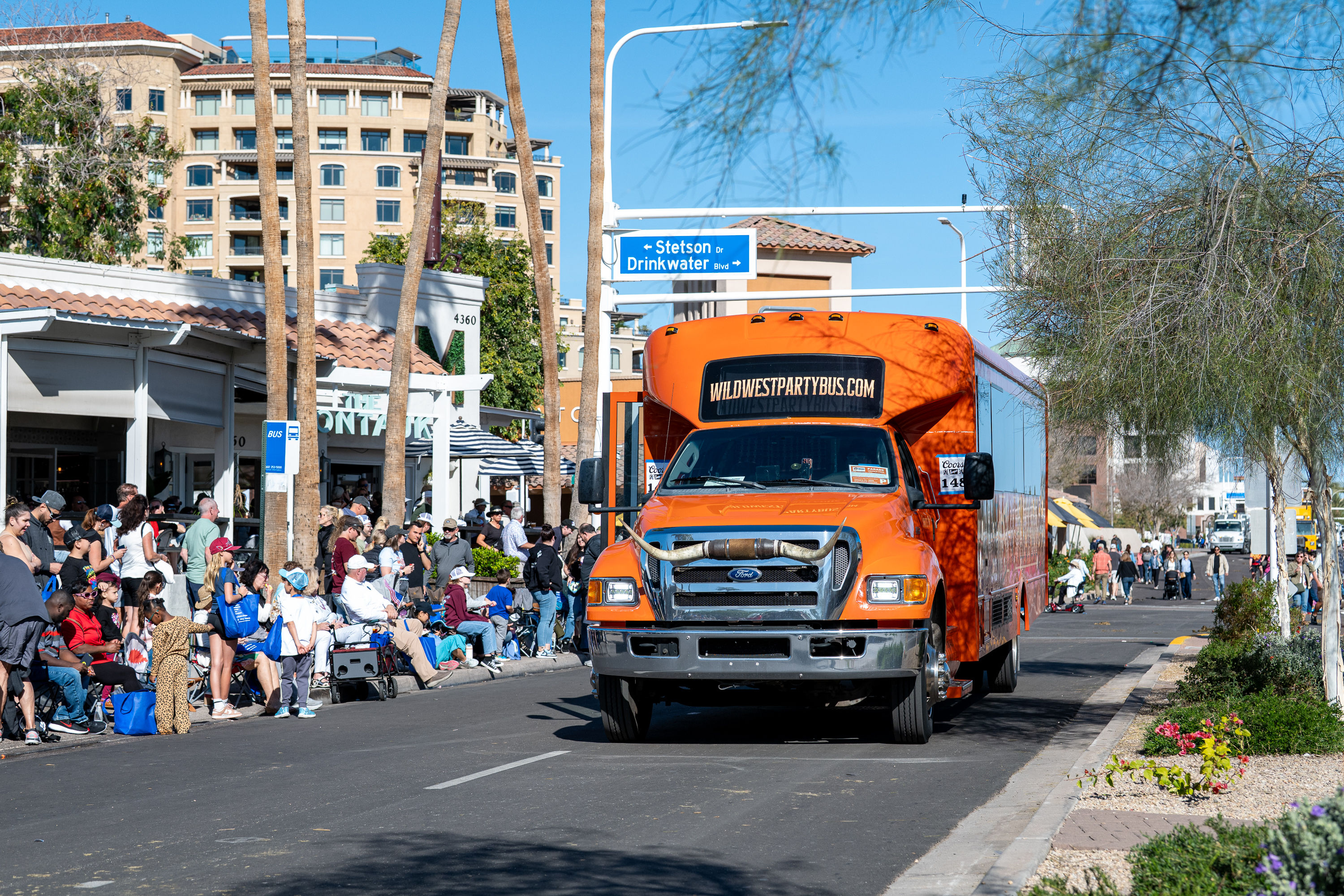 Orange party bus driving down a sunny downtown shopping street lined with palm trees, crowds seated on the sidewalk watching a parade.