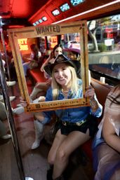 Smiling woman in a cowboy hat and denim shirt holds a 'Wanted' photo frame aboard a neon-lit, cowboy-themed party bus, surrounded by friends in boots and hats.