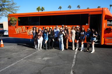 Cheerful group in cowgirl outfits and boots posing in front of an orange western-themed party bus in a sunny parking lot with palm trees.