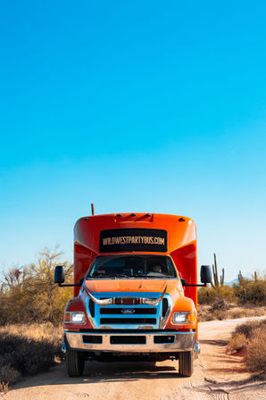 Front-facing bright orange party bus on a sandy desert road, framed by saguaro cacti and scrub under a vivid blue southwestern sky.