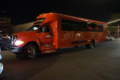 Orange Western-themed party bus with longhorn hood ornament parked on a downtown street at night, string lights and a palm tree in the background