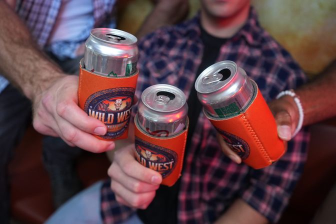 Three friends toasting open canned drinks in orange koozies, casual indoor bar/party vibe with plaid shirts