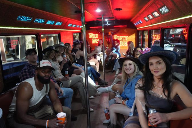 Group of young adults in cowboy hats and casual outfits holding drinks and smiling inside a neon-lit, wood-paneled party bus with city street visible outside.
