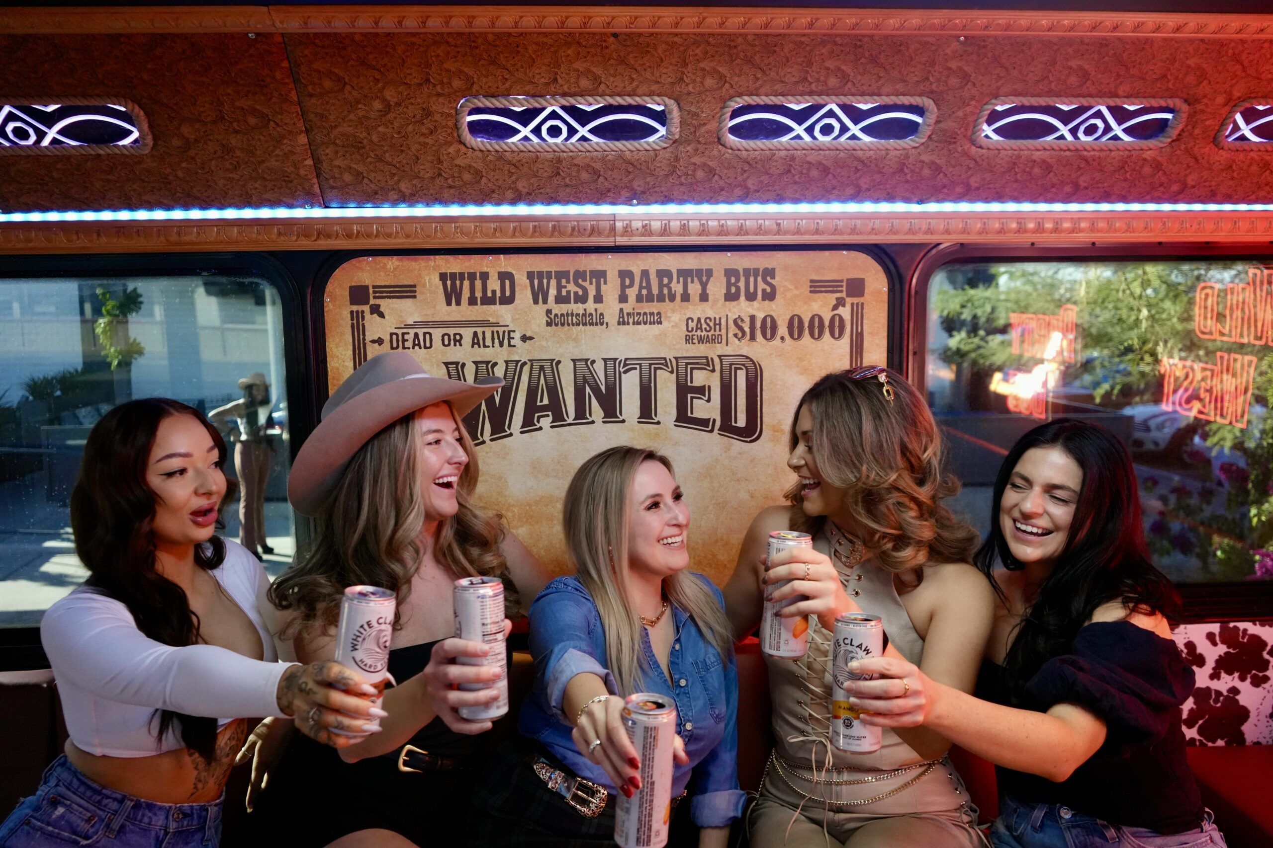 Five friends laughing and toasting canned drinks inside a Western-themed party bus with a large "WANTED" poster backdrop, cowgirl hat and colorful LED lighting.