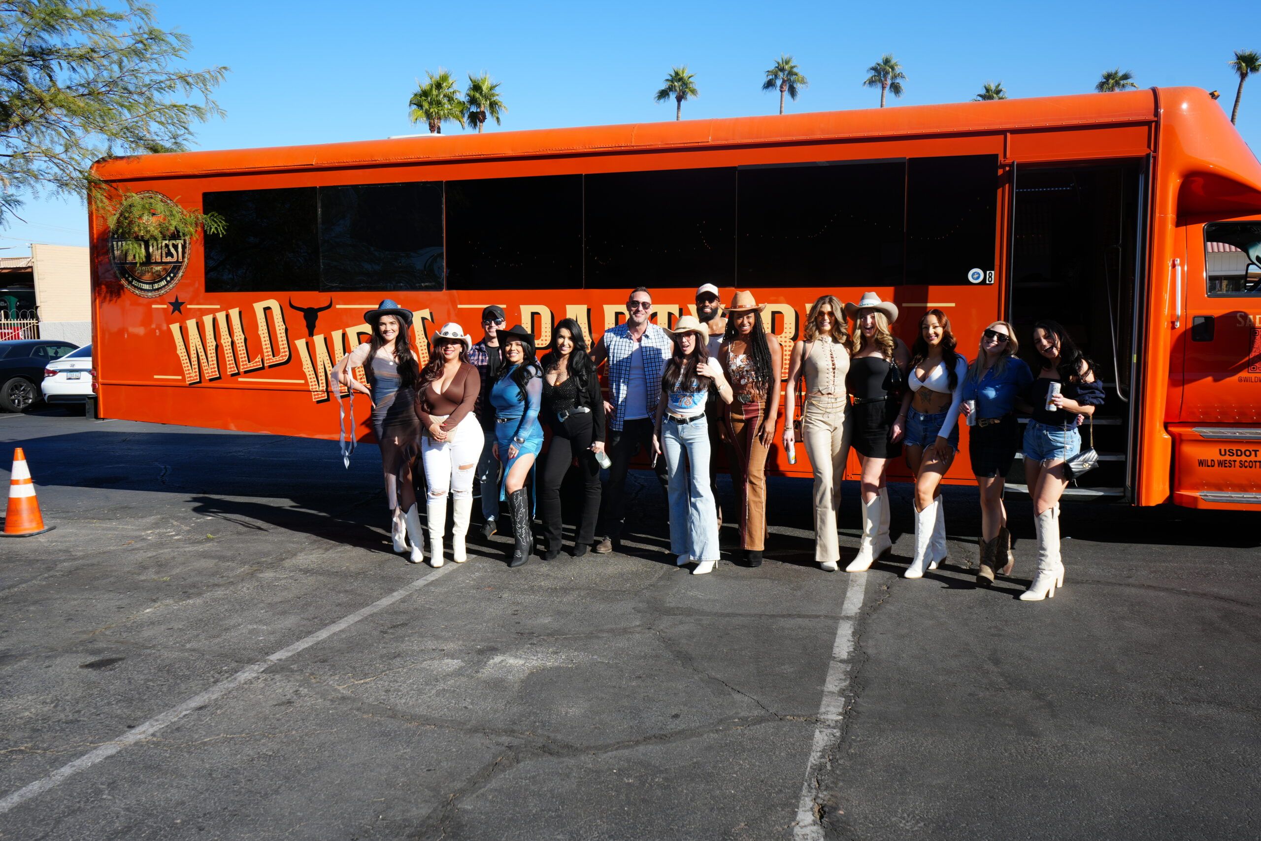 Energetic group of people in cowboy hats and boots posing in front of a bright orange party bus in a sunny parking lot with palm trees.