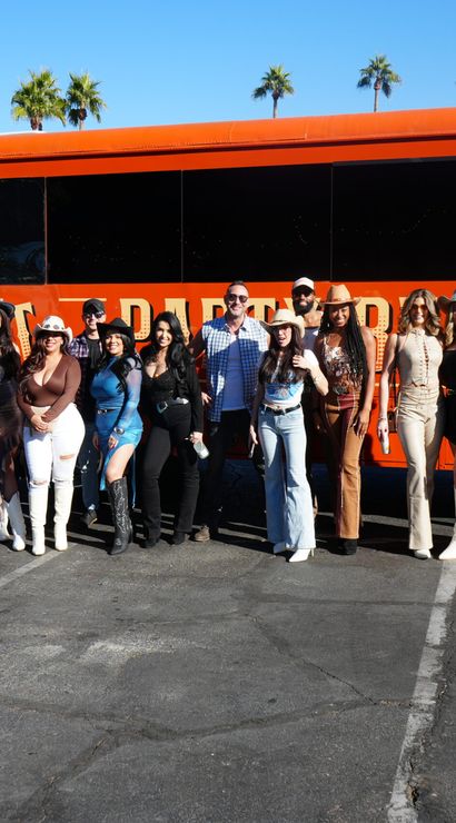 Energetic group of people in cowboy hats and boots posing in front of a bright orange party bus in a sunny parking lot with palm trees.
