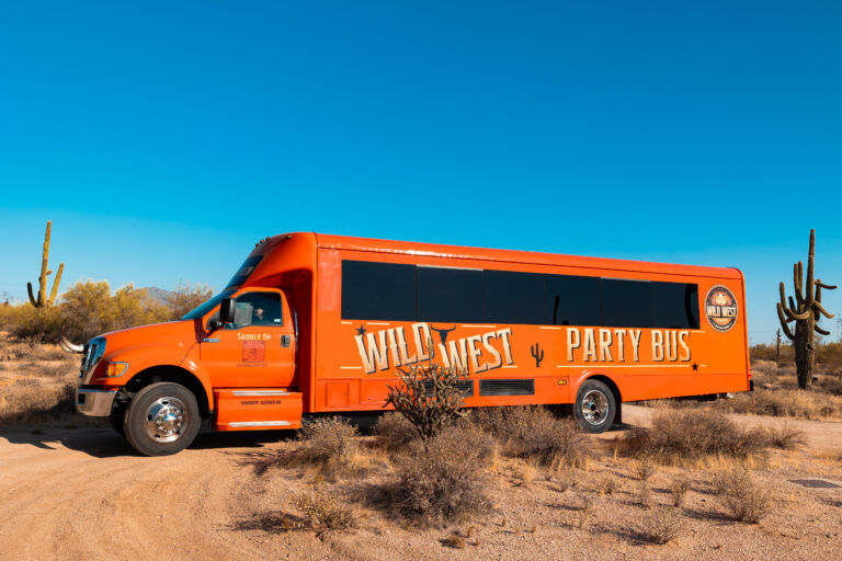 Bright orange party bus with western-style lettering parked on a sandy trail in a sunny Southwestern desert, surrounded by saguaro cacti and scrub under a clear blue sky.