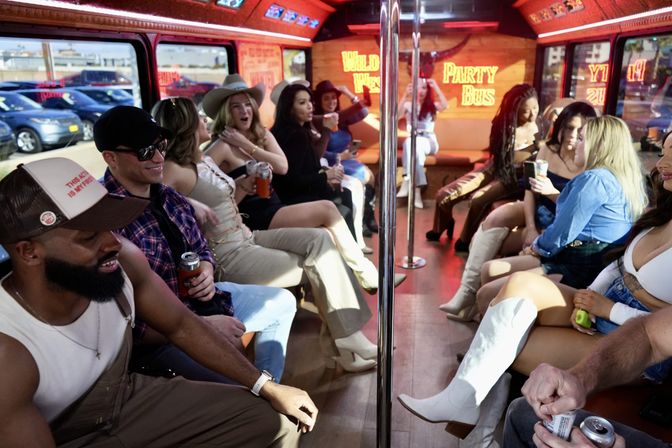 Neon-lit party bus interior with a group of friends seated on bench seats around a chrome dance pole, laughing, dancing and sipping drinks during a daytime city ride.