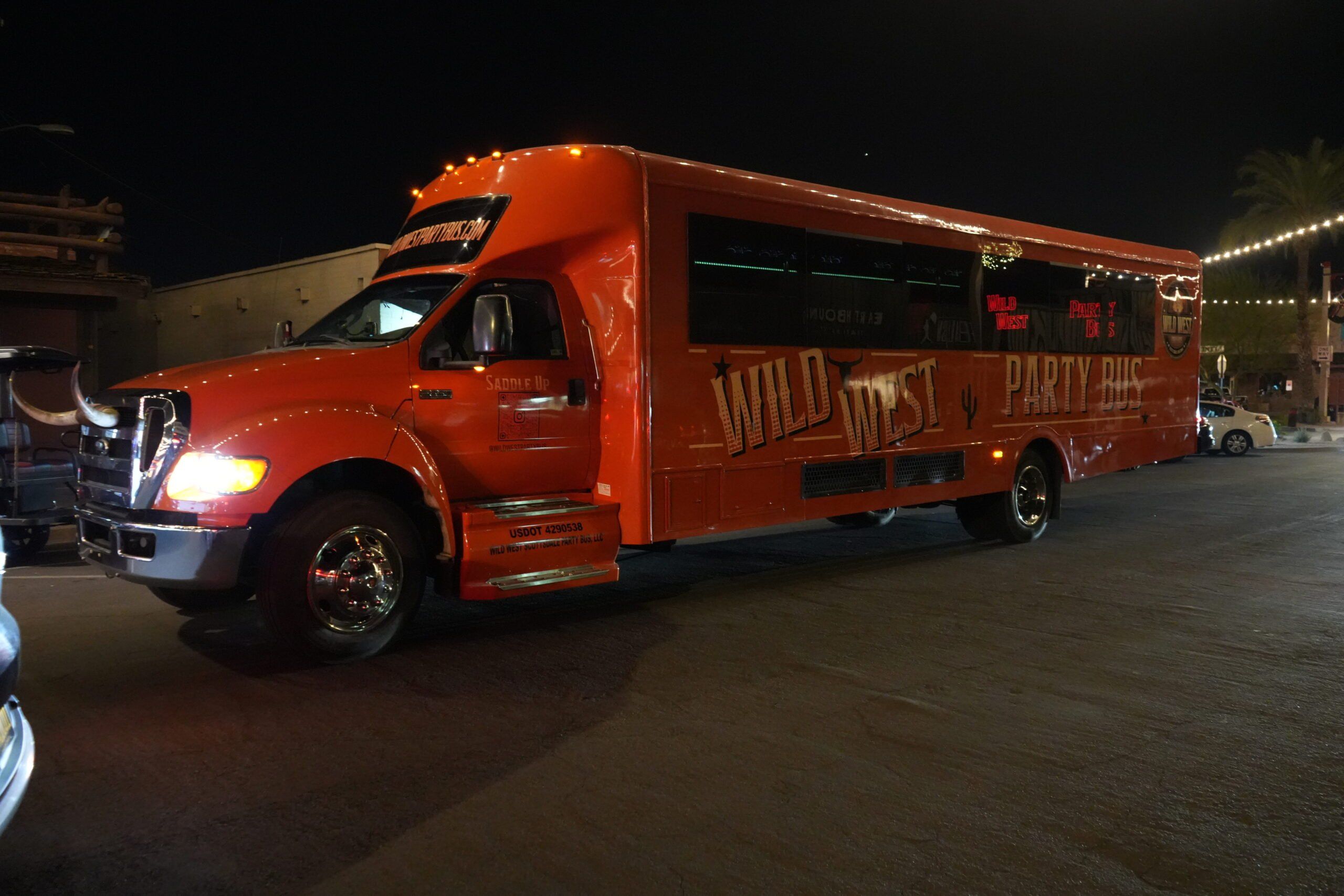 Orange Western-themed party bus with longhorn hood ornament and illuminated windows parked on a downtown street at night under string lights.