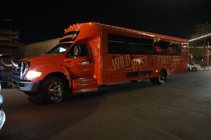Orange Western-themed party bus with longhorn hood ornament and illuminated windows parked on a downtown street at night under string lights.