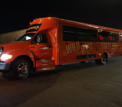 Orange Western-themed party bus with longhorn hood ornament and illuminated windows parked on a downtown street at night under string lights.