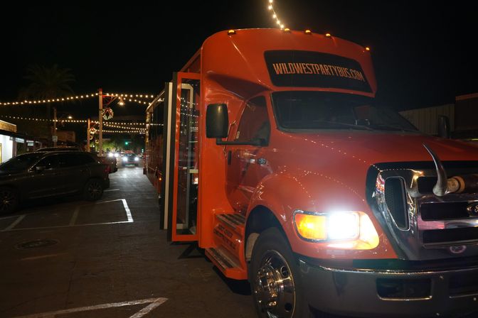 Bright orange party bus parked on a downtown street at night under hanging string lights, headlights glowing and decorative longhorn on the grille, with nearby cars and a palm tree