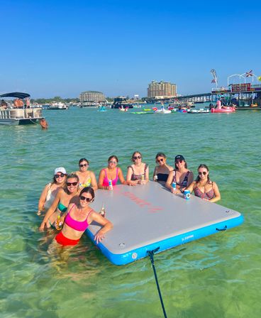 Group of friends relaxing on a large inflatable dock in shallow turquoise bay, holding drinks amid boats, colorful floaties, a nearby pier and waterfront condos under a clear blue sky