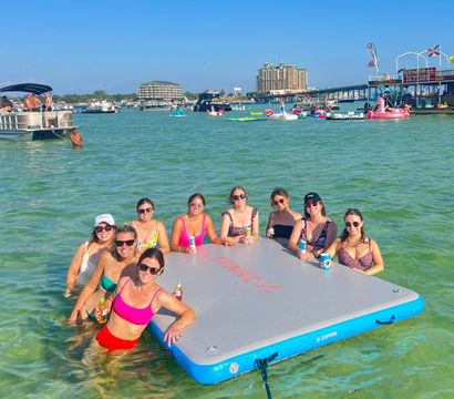 Group of friends relaxing on a large inflatable dock in shallow turquoise bay, holding drinks amid boats, colorful floaties, a nearby pier and waterfront condos under a clear blue sky