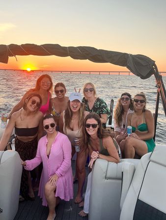 Group of women posing on a pontoon boat at sunset, holding drinks and wearing swimsuits and sunglasses with ocean and a long bridge on the horizon.