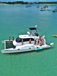 White catamaran anchored in shallow turquoise bay with a group of people in swimsuits socializing on deck; clear water, other boats and a sandy, tree-lined shore in the background.