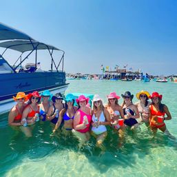 Group of women in colorful swimsuits and cowboy hats smiling and holding drinks waist-deep in clear turquoise water beside a pontoon boat at a lively sandbar party with inflatables and boats in the background.