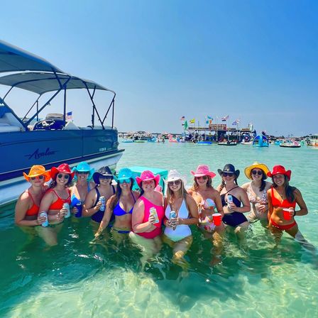 Group of women in colorful swimsuits and cowboy hats smiling and holding drinks waist-deep in clear turquoise water beside a pontoon boat at a lively sandbar party with inflatables and boats in the background.