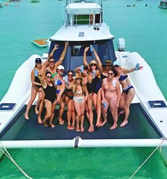 Group of friends in swimsuits cheering with canned drinks on the bow of a white yacht over clear turquoise tropical water