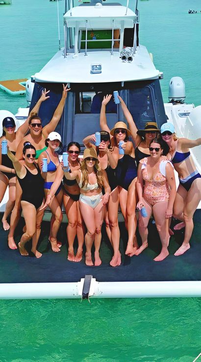 Group of friends in swimsuits cheering with canned drinks on the bow of a white yacht over clear turquoise tropical water