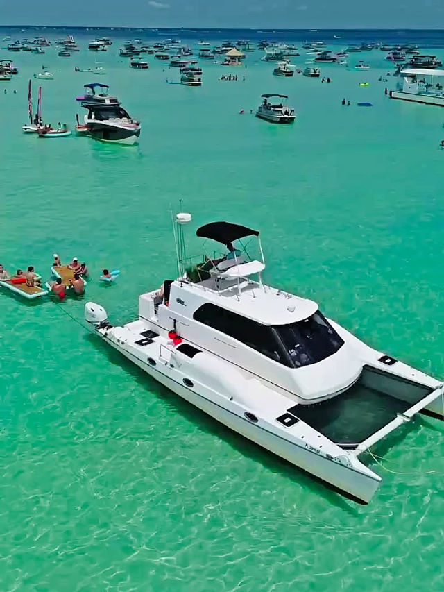 Aerial view of a white catamaran anchored in shallow turquoise tropical water, surrounded by dozens of boats and people on inflatables enjoying a sunny day