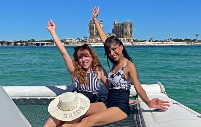 Two smiling women in black-and-white swimsuits celebrating on a boat deck over clear turquoise coastal water, a straw sunhat embroidered "BRIDE" on a lap, and beachfront resort towers on the horizon.