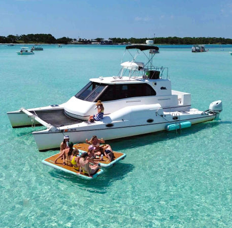 White catamaran anchored in shallow turquoise lagoon, group of friends relaxing on a floating mat on a sunny day
