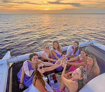 Smiling group of friends in colorful dresses and sashes toasting with drinks while seated on a catamaran’s net during a golden ocean sunset cruise.