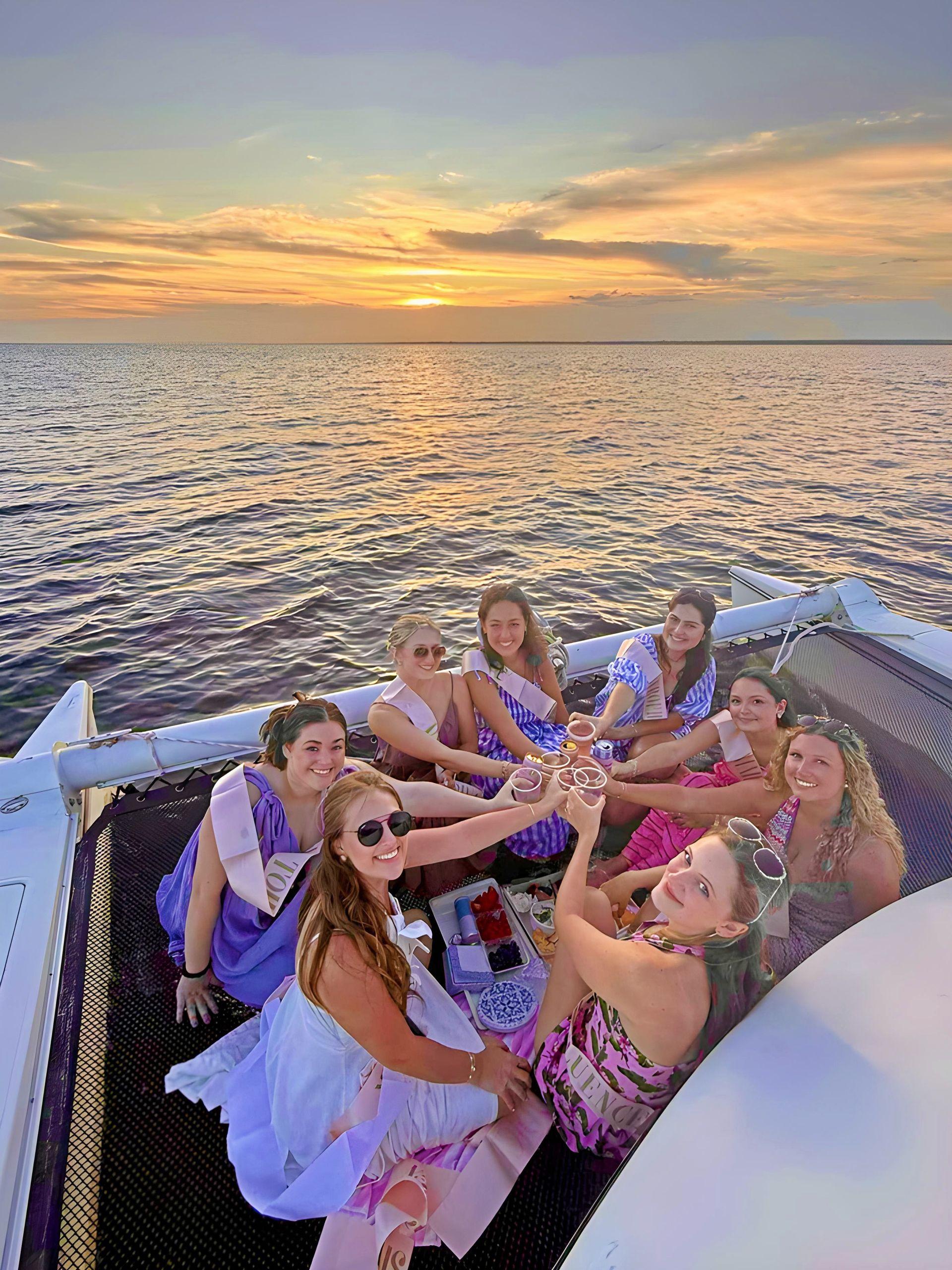 Smiling group of friends in colorful dresses and sashes toasting with drinks while seated on a catamaran’s net during a golden ocean sunset cruise.