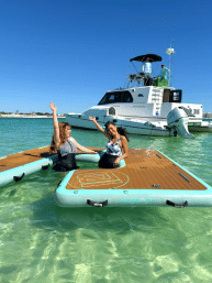 Two women waving and smiling on inflatable wooden-look floating docks in clear turquoise water beside a white motor yacht on a sunny coastal day