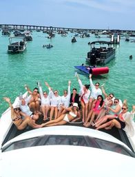 Group of friends partying on the bow of a white yacht in crystal-clear turquoise water, surrounded by dozens of anchored boats and a distant bridge on a sunny summer day.