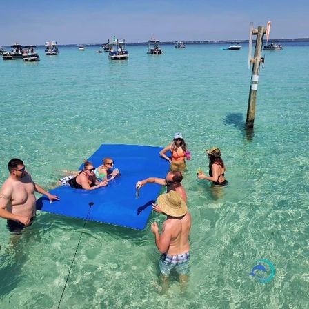 Group of friends relaxing on a bright blue floating mat in shallow turquoise beach water near anchored boats and a wooden marker — summer sandbar scene.