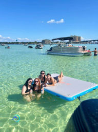 Group of four friends relaxing in clear shallow turquoise bay—two standing in waist-deep water and two lounging on a rectangular inflatable mat, with pontoon boats, a bridge and coastal buildings under a bright blue sky.