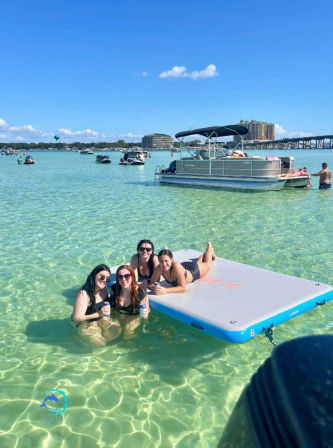 Group of four friends relaxing in clear shallow turquoise bay—two standing in waist-deep water and two lounging on a rectangular inflatable mat, with pontoon boats, a bridge and coastal buildings under a bright blue sky.