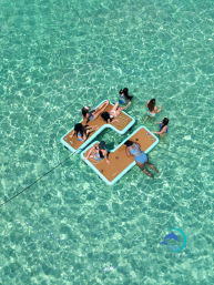 Aerial view of people in colorful swimsuits lounging on linked floating platforms in crystal-clear turquoise tropical water, enjoying a sun-soaked shallow sea.